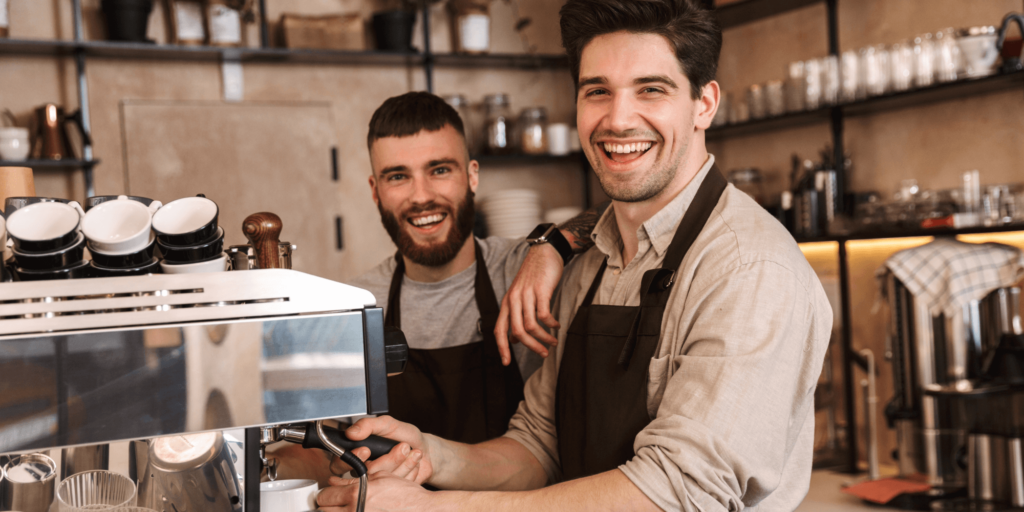 Smiling male baristas in aprons preparing coffee behind a modern espresso machine in a lively café setting, highlighting hospitality and teamwork.