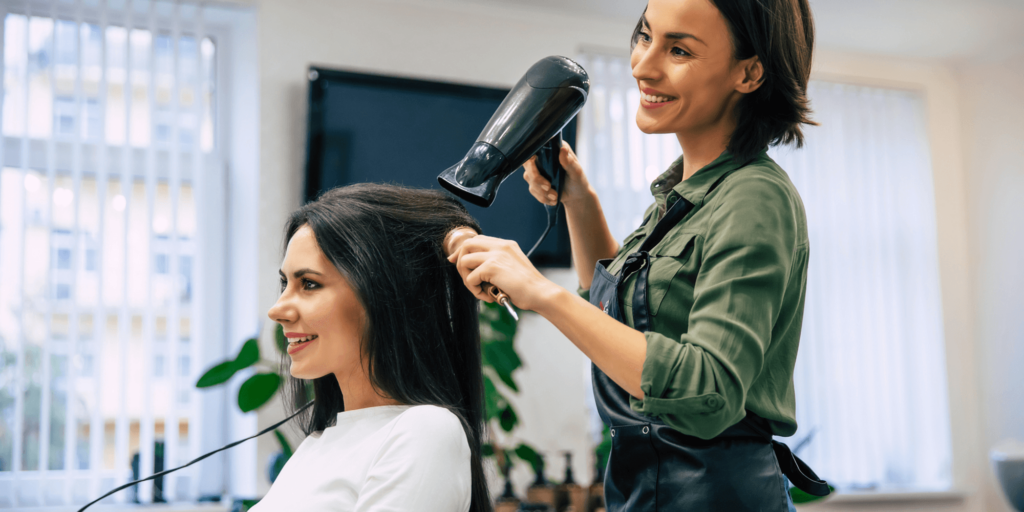 A smiling female hairdresser using a hairdryer and round brush to style a female client’s hair in a bright, modern salon setting.