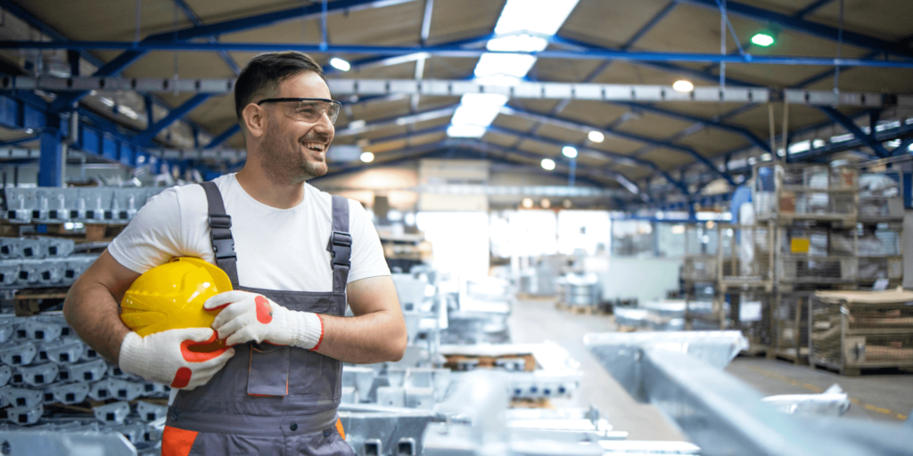 Smiling factory worker in gloves holding a yellow hard hat inside a manufacturing facility.